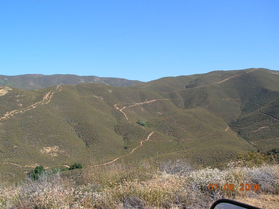 Dirt road winds through a dry, brush-covered mountain landscape under a clear blue sky. A date stamp reads 07.08.2006
