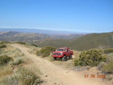 1985 Ford Bronco - A red pickup truck is parked on a dirt road in a hilly, arid landscape under a clear blue sky.