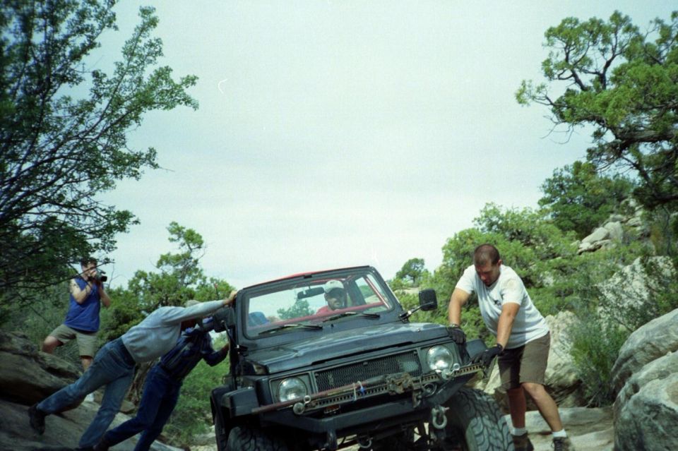 Green open-top Jeep on rocky trail with spotters bracing it as it tips back on a ledge