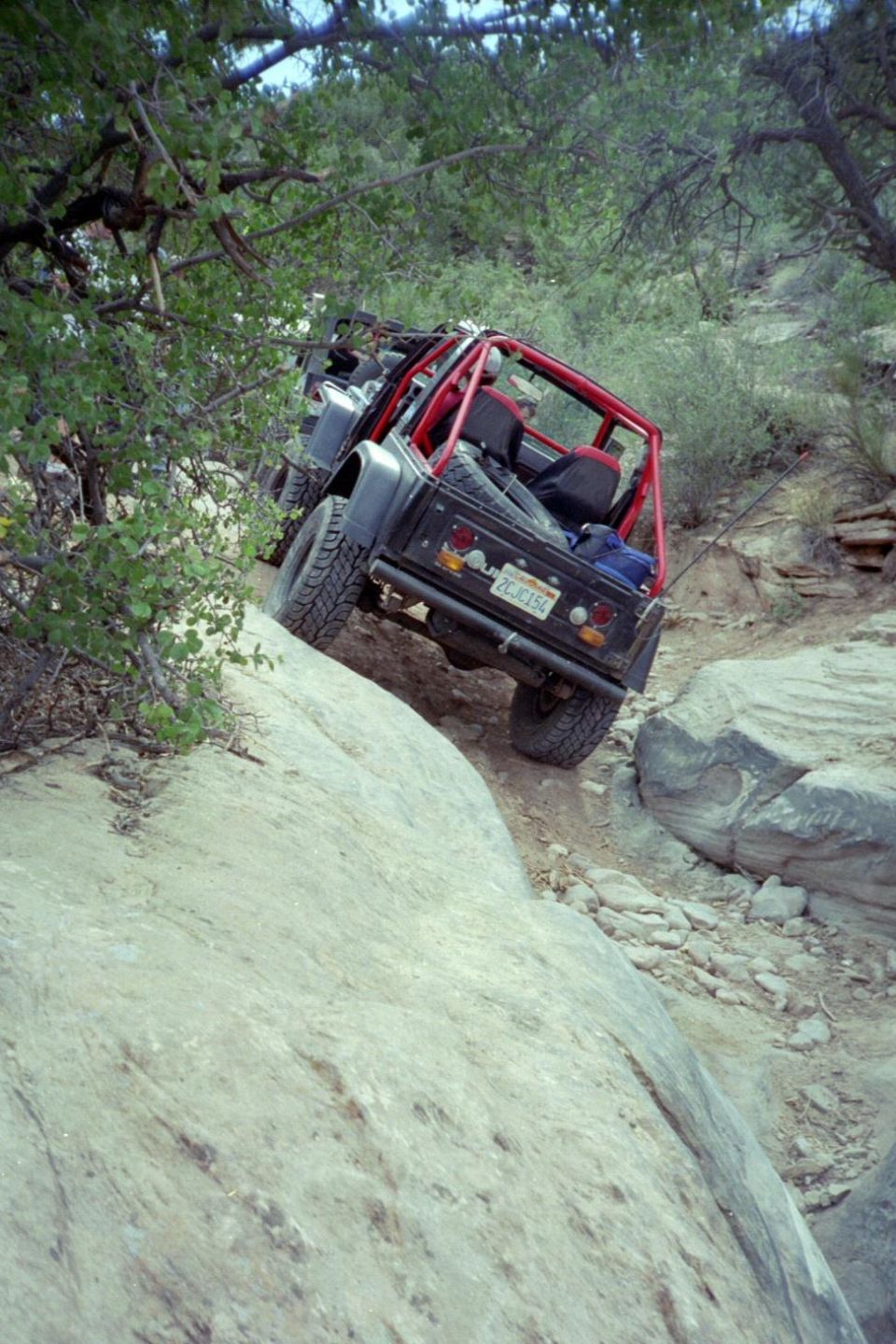 Grey open-top 4x4 with red roll cage descending an off-camber slickrock ledge