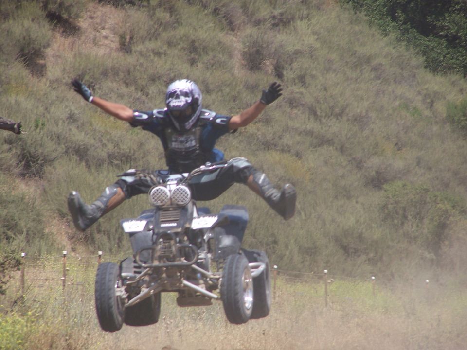 Rider doing airborne trick on dark blue sport quad ATV over dusty hillside trail
