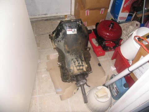 1978 Ford Bronco - Transmission and torque converter removed from a vehicle, resting on cardboard with surrounding storage items.