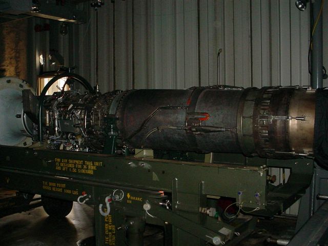 Jet engine mounted on a green stand with various hoses and wires attached. Text is visible on the stand
