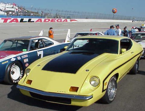 1972 Ford Ranchero - A yellow 1972 Ford Ranchero is parked at a racetrack, with other race cars and spectators in the background.