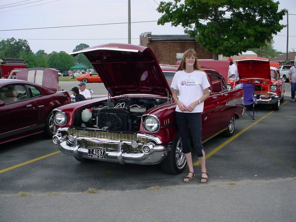 Woman standing next to a red 1957 Chevrolet Bel Air with hood open. Another red classic car is parked behind it