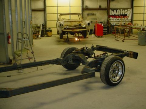 1972 Ford Ranchero - A car chassis with chrome wheels and whitewall tires is the focus in a garage, with another vehicle in the background.