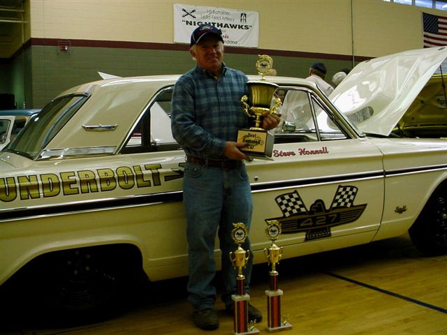Man holding trophies next to a 1964 Ford Thunderbolt. Car has 'Thunderbolt' and '427' graphics