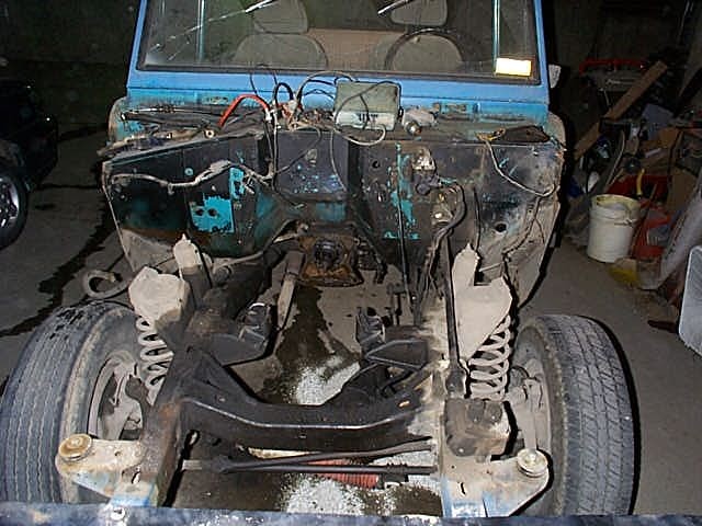 Front of a teal truck stripped down in a garage, engine removed, front axle and coil springs exposed