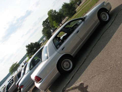 2003 Ford Crown Victoria - Several silver sedans are parked in a row next to a body of water on a sunny day.