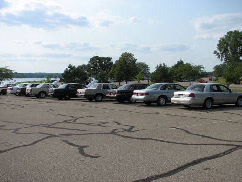 2003 Ford Crown Victoria - Several Ford Crown Victorias are parked in a lot near a body of water under a cloudy sky.
