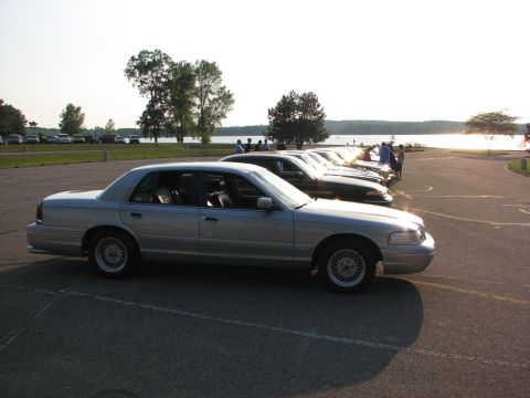 2003 Ford Crown Victoria - Several cars are parked in a lot near a body of water on a sunny day.
