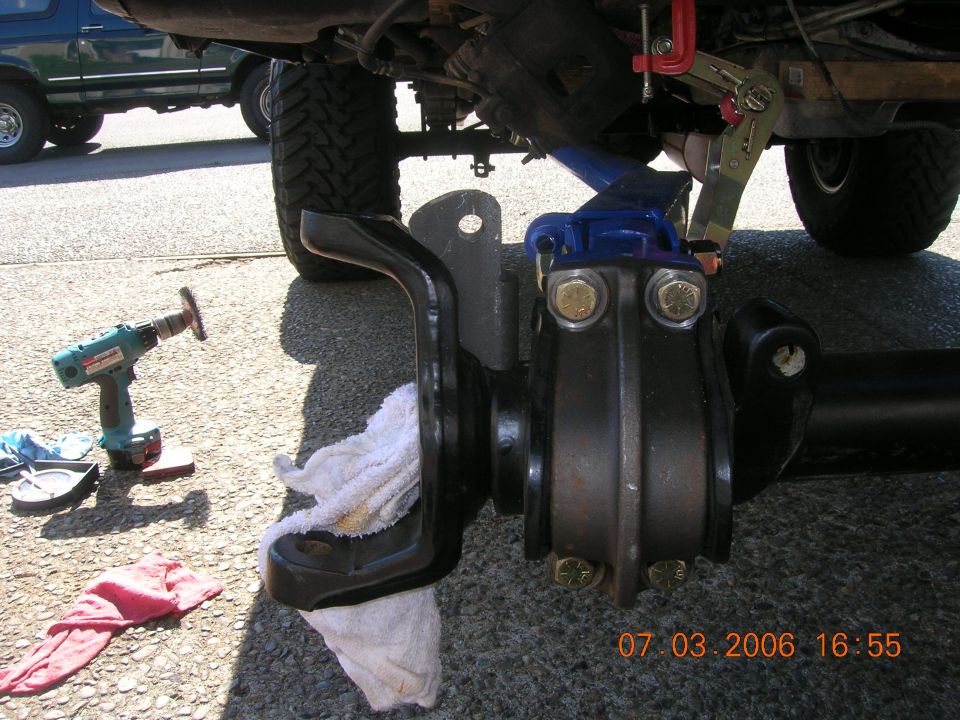 Black-painted solid axle end and bracket on the ground under a lifted truck during suspension work