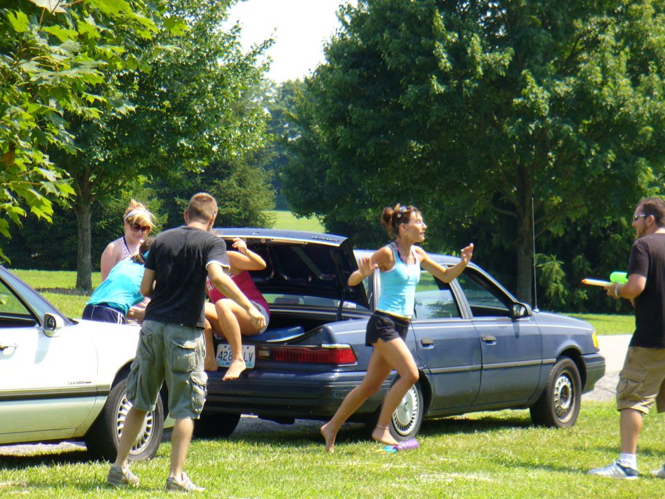 Blue sedan with open trunk, people interacting, one person in trunk, another running. License plate 426 LV