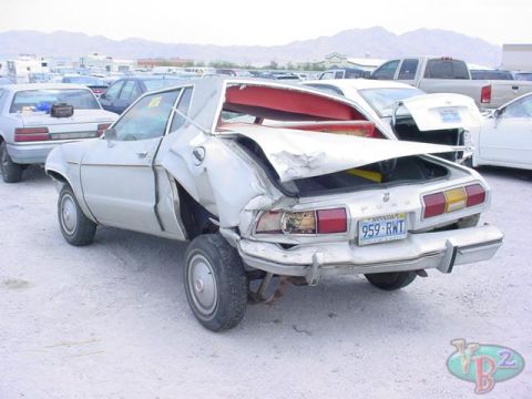 1978 Ford Mustang II - This severely damaged sedan is parked in a lot with other vehicles under a bright sky.