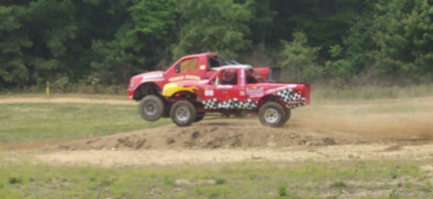 Two red monster trucks mid-air over a dirt jump
