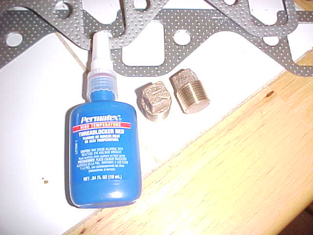 Permatex red threadlocker bottle with two brass pipe plugs and exhaust gaskets on a workbench