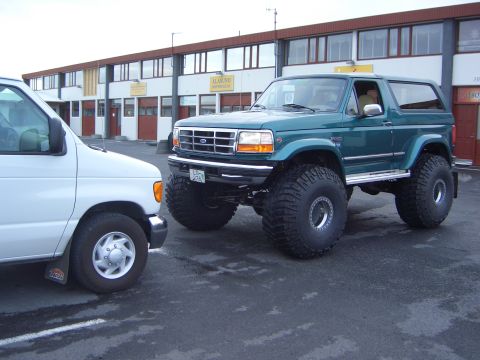 1996 Ford Bronco - A lifted 1996 Ford Bronco with large off-road tires is parked next to a white van in front of a commercial building.