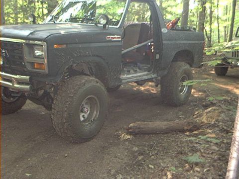 1986 Ford Bronco - A modified Ford Bronco sits in a dirt clearing, its door ajar, with another vehicle visible in the background.