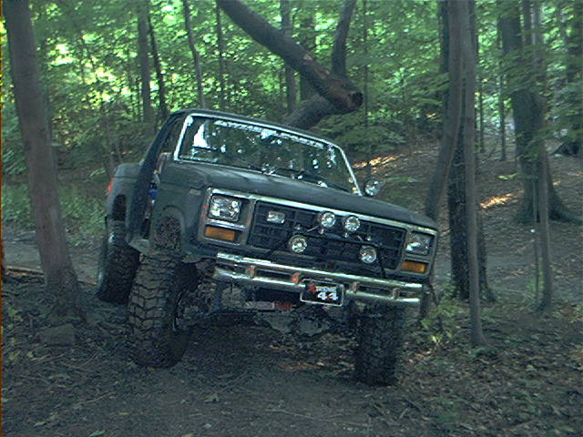 Black SUV with off-road tires driving through a wooded area. Has extra lights on the front