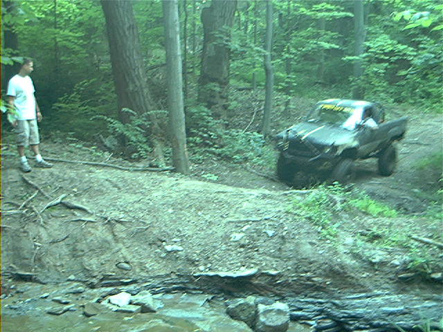 Muddy off-road truck with yellow text on windshield, driving on a dirt path in a forest