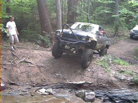 1986 Ford Bronco - A heavily modified off-road truck navigates a steep dirt incline in a wooded area, with a white Jeep visible further up the path.