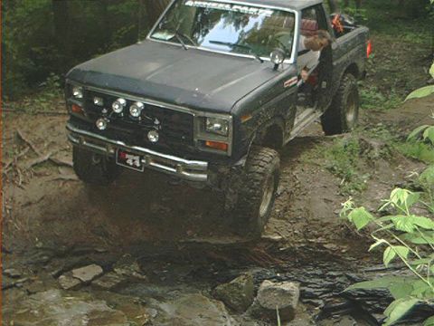 1986 Ford Bronco - This dark-colored SUV, likely from the early 1980s, navigates a challenging muddy terrain with water flowing across its path.