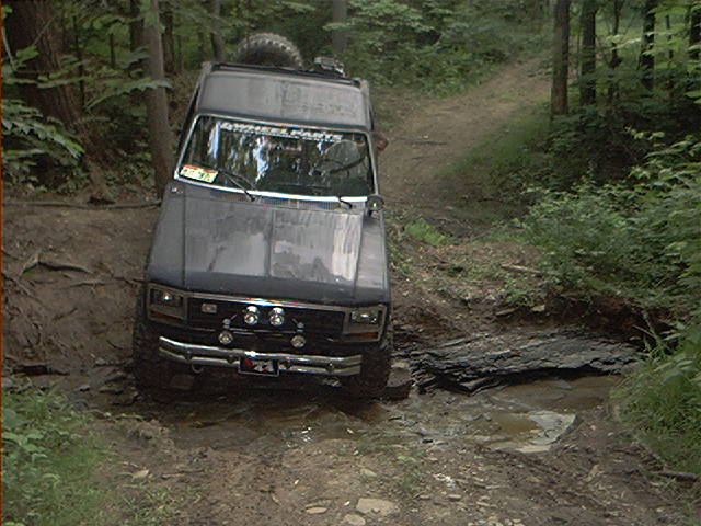 Dark gray Ford Bronco navigating a muddy, wooded trail; spare tire mounted on roof rack