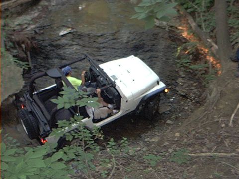 1986 Ford Bronco - A white Jeep navigates a challenging off-road trail, splashing through water and over rocks.
