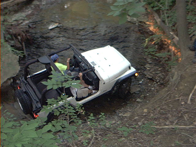 White Jeep Wrangler driving through a rocky stream bed with trees on either side. Two people are inside