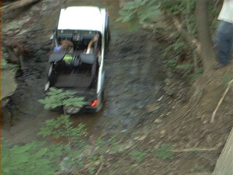 1986 Ford Bronco - A white off-road SUV navigates a muddy stream with its top removed.