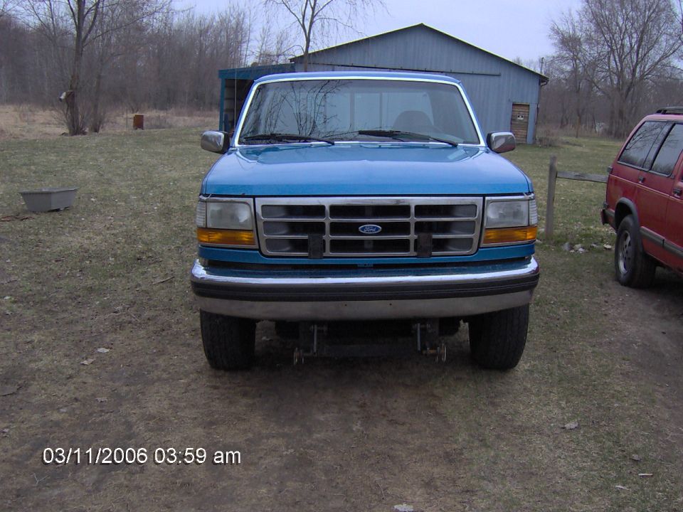 Front view of blue Ford F-150 with chrome grille and plow mount, parked by a gray barn.