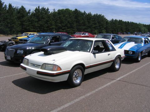 1989 Ford Mustang - A white 1989 Ford Mustang coupe with a prominent hood scoop and racing stripes is parked in a line with other vehicles.