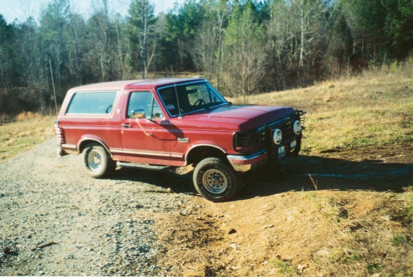 Red lifted Ford Bronco SUV with brush guard and off-road tires on a gravel road near woods