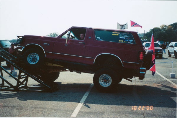 Lifted red Ford Bronco climbing a ramp at an event, Confederate and club flags flying behind