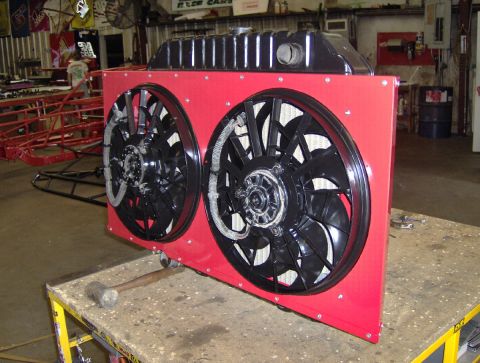 1978 Ford Bronco - A radiator with a black top tank sits on a shop workbench fitted with a custom red sheet-metal shroud carrying twin black electric cooling…