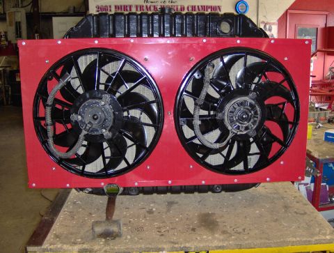 1978 Ford Bronco - Head-on view of a completed radiator and dual-electric-fan assembly standing on a shop bench, the two black fans bolted into a red riveted…