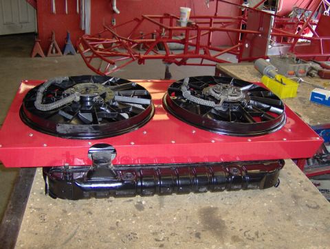 1978 Ford Bronco - Top-down view on a shop bench of a radiator with a gloss-black top tank and filler neck, topped by a custom red sheet-metal shroud holding…