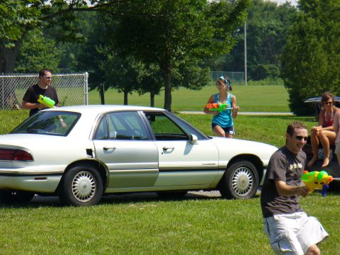 1991 Ford Probe - A group of people with water guns gathered around a white sedan in a park setting.