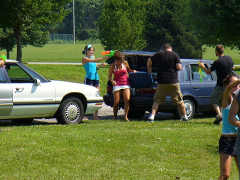 1991 Ford Probe - A group of people engage in a water gun fight next to two cars parked on a grassy area.