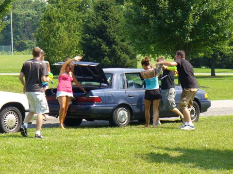 1991 Ford Probe - People interact around a blue sedan with its trunk open in a grassy park area.