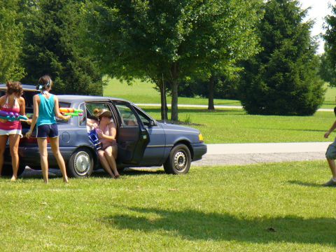 1991 Ford Probe - People are gathered around a blue sedan with water guns, ready for a water fight on a sunny day.