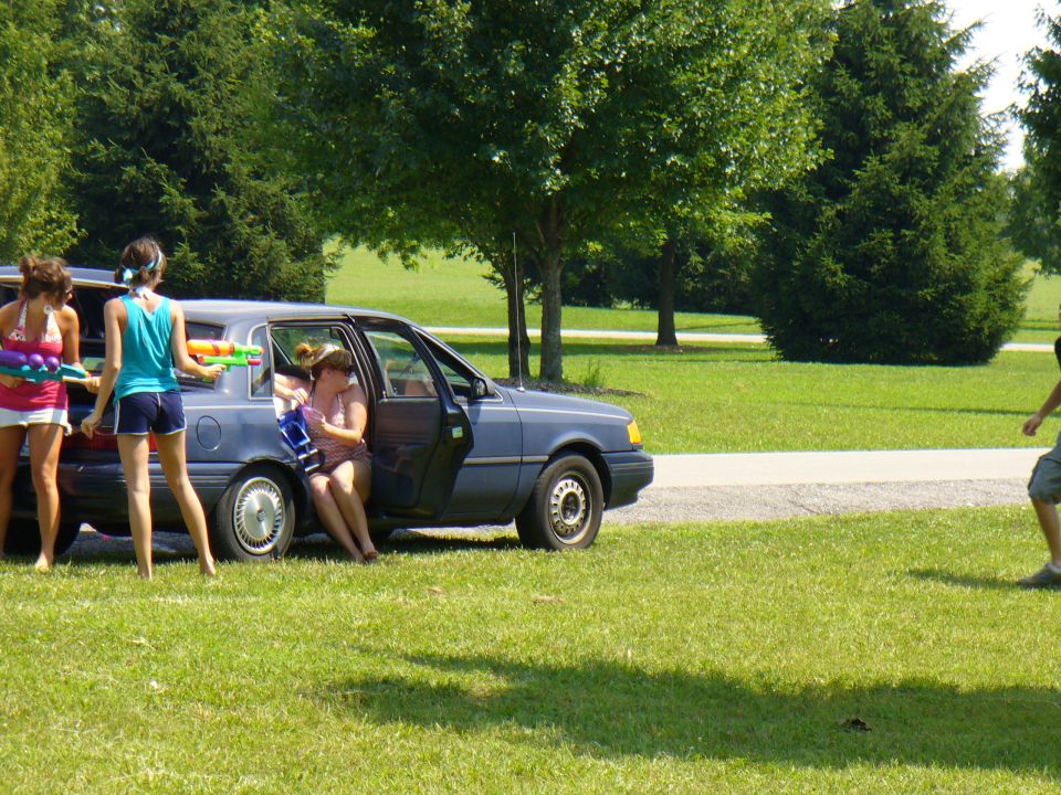 Blue sedan with open doors, people with water guns in and around it, on a grassy area