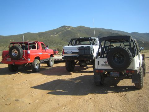 1990 Ford Bronco - red lifted truck on a trailer, a white truck, and a white Suzuki 4x4 parked on a dirt staging area before green hills.