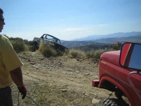 1990 Ford Bronco - man in yellow stands beside a red rig watching a lifted truck navigate a rocky hilltop trail with mountains beyond.