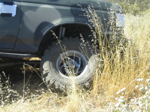 1990 Ford Bronco - Front corner of a dark lifted truck mired in tall dry grass, showing a large mud-terrain off-road tire and long shock.