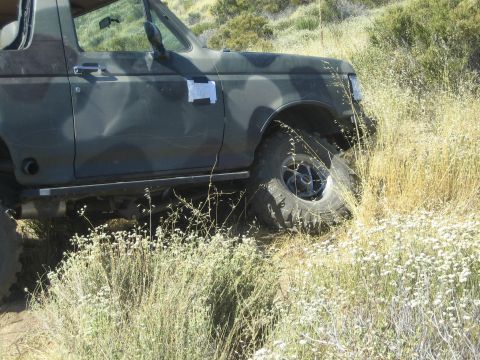 1990 Ford Bronco - dark camo-painted Ford truck high-centered in dry brush and wildflowers off the trail on a hillside.