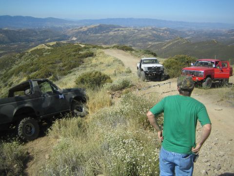 1990 Ford Bronco - man in a green shirt watches a black, white, and red truck staged along a ridge-top dirt trail over rolling green hills.