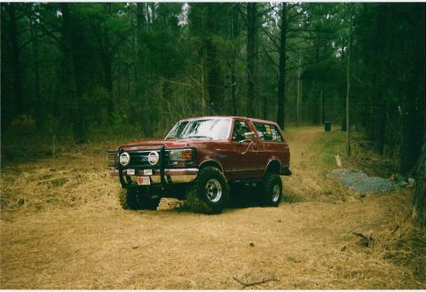 1991 Ford Bronco - A lifted red Ford Bronco sits on a dirt path surrounded by trees.
