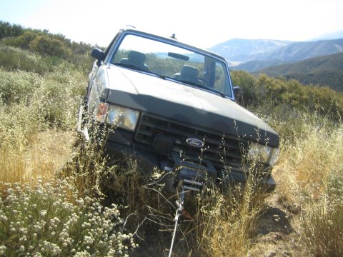 1990 Ford Bronco - dark soft-top Ford truck with a front winch high-centered in dry brush on a hillside, mountains in the distance.