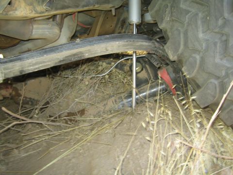 1990 Ford Bronco - Underbody view of a lifted truck showing a long shock absorber, axle components and a large off-road tire amid dirt and dry grass.